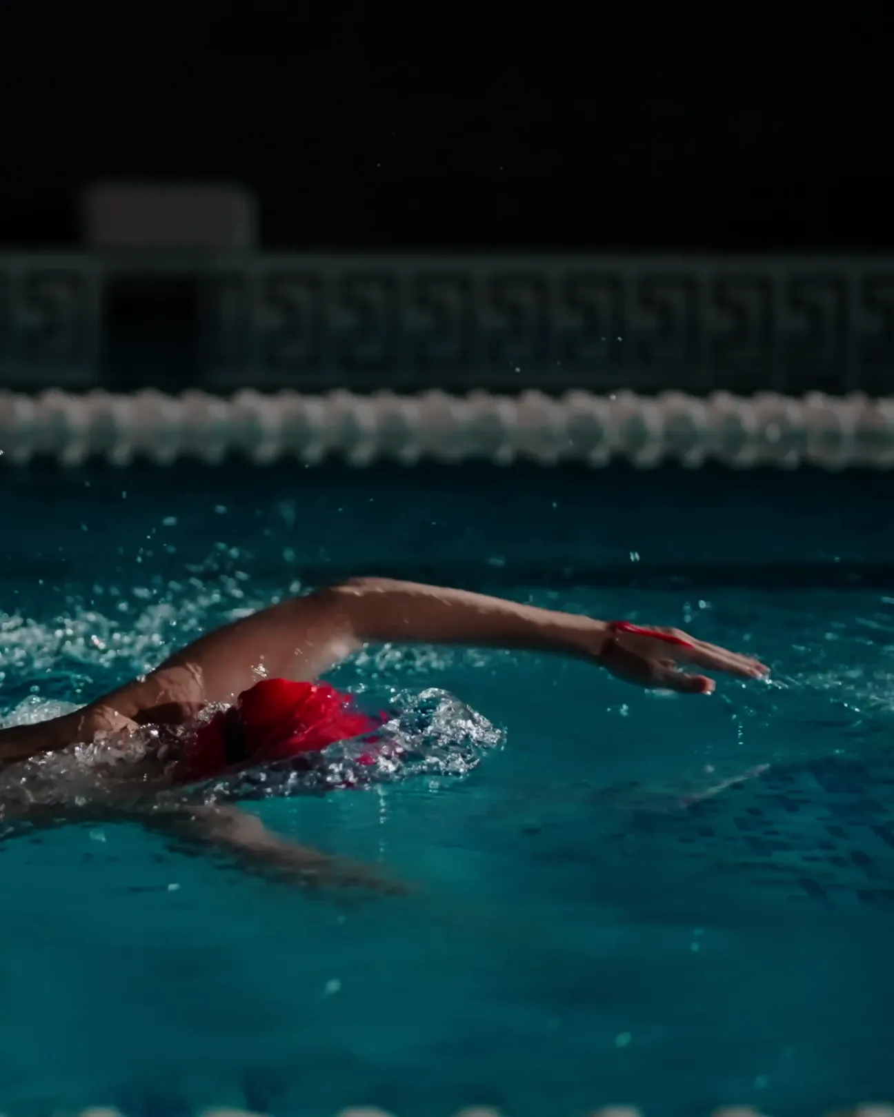La nuit, une nageuse en bonnet rouge nage le crawl d’un geste ample et régulier, le bras allongé loin devant elle au-dessus de l’eau.